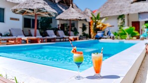 Un área junto a la piscina con sillones de descanso y sombrillas de paja, dos cócteles coloridos y una piscina azul cristalina.