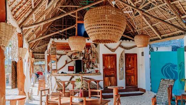 Un restaurante junto a la playa con mobiliario de madera y techo de paja.