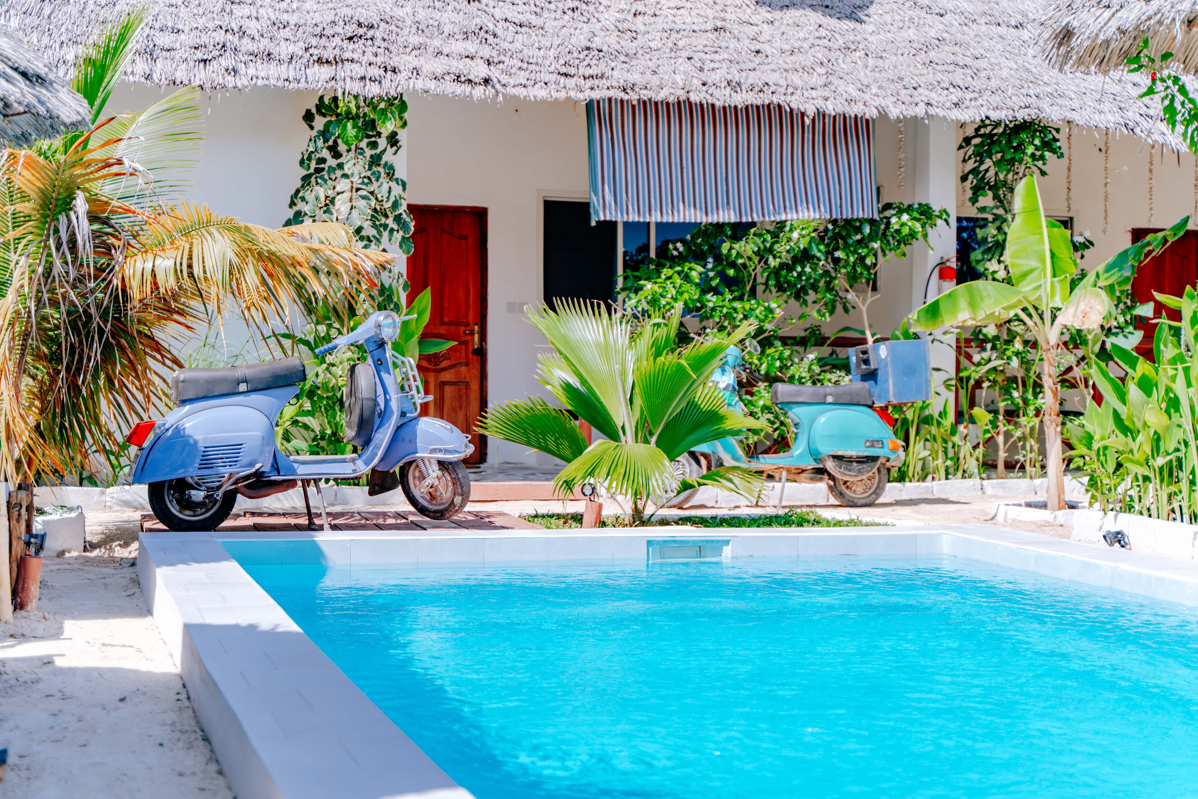 A pool area with a blue scooter parked by the poolside, surrounded by tropical plants and a thatched roof structure.