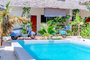A pool area with a blue scooter parked by the poolside, surrounded by tropical plants and a thatched roof structure. - Simba Apartment Hotel (Bwejuu)