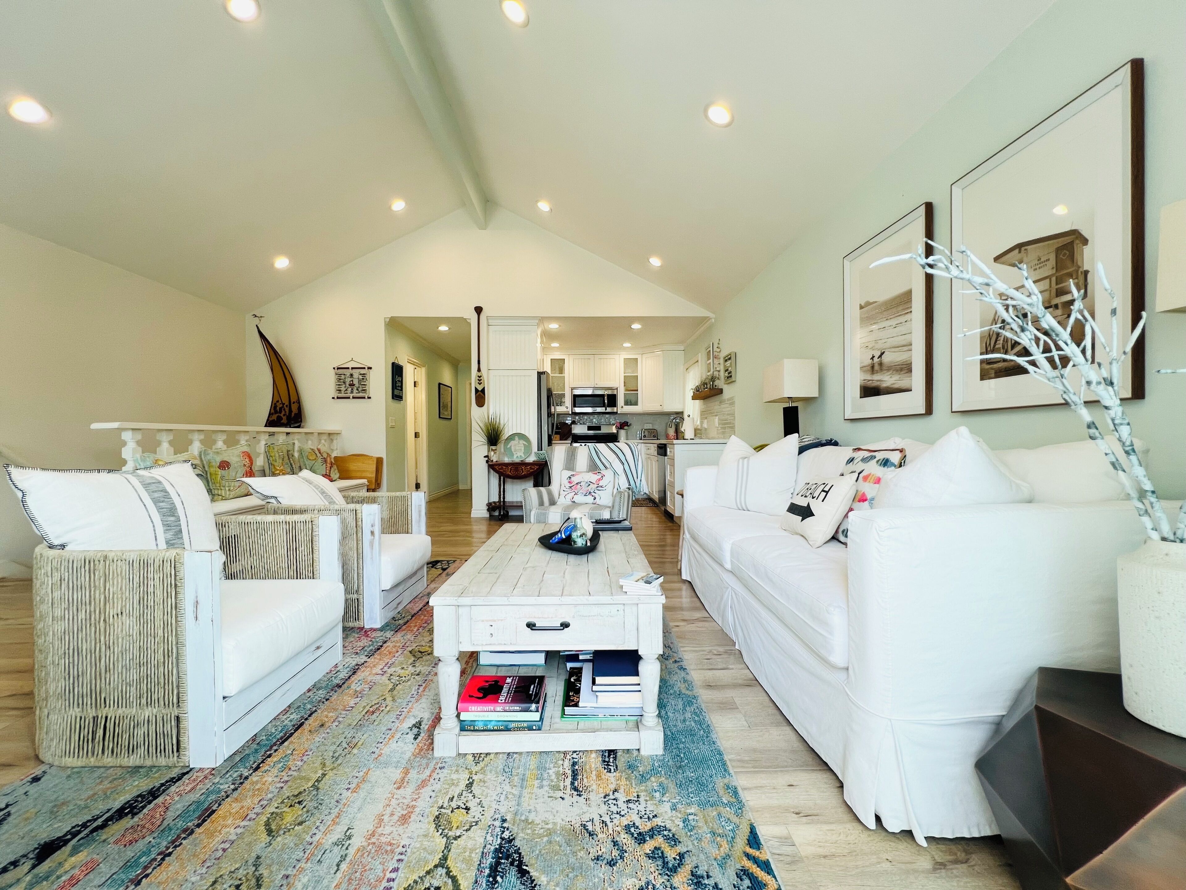 A spacious living room with white furniture, a patterned rug, and a kitchen in the background.