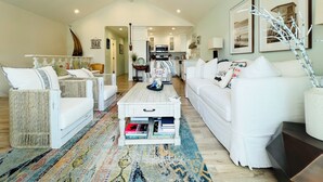 Un salón amplio con muebles blancos, una alfombra estampada y una cocina al fondo.