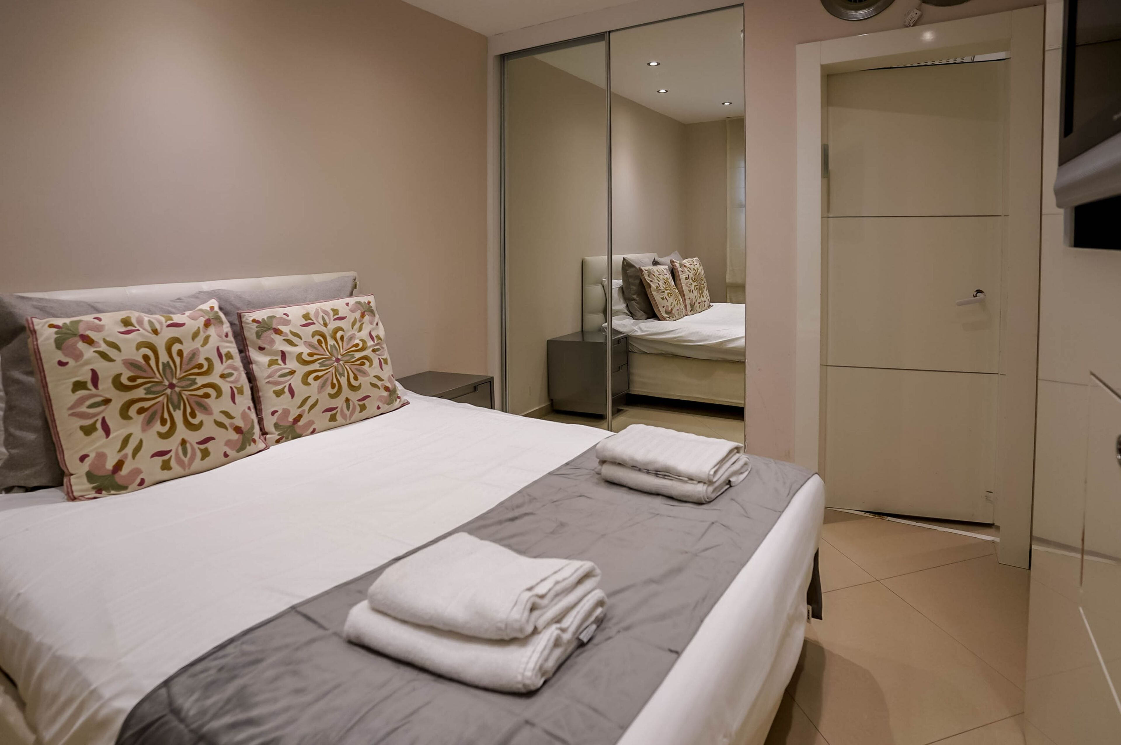 Una cama bien hecha con almohadas florales, una colcha gris y toallas dobladas. Hay un armario con espejo y un televisor montado en la pared.