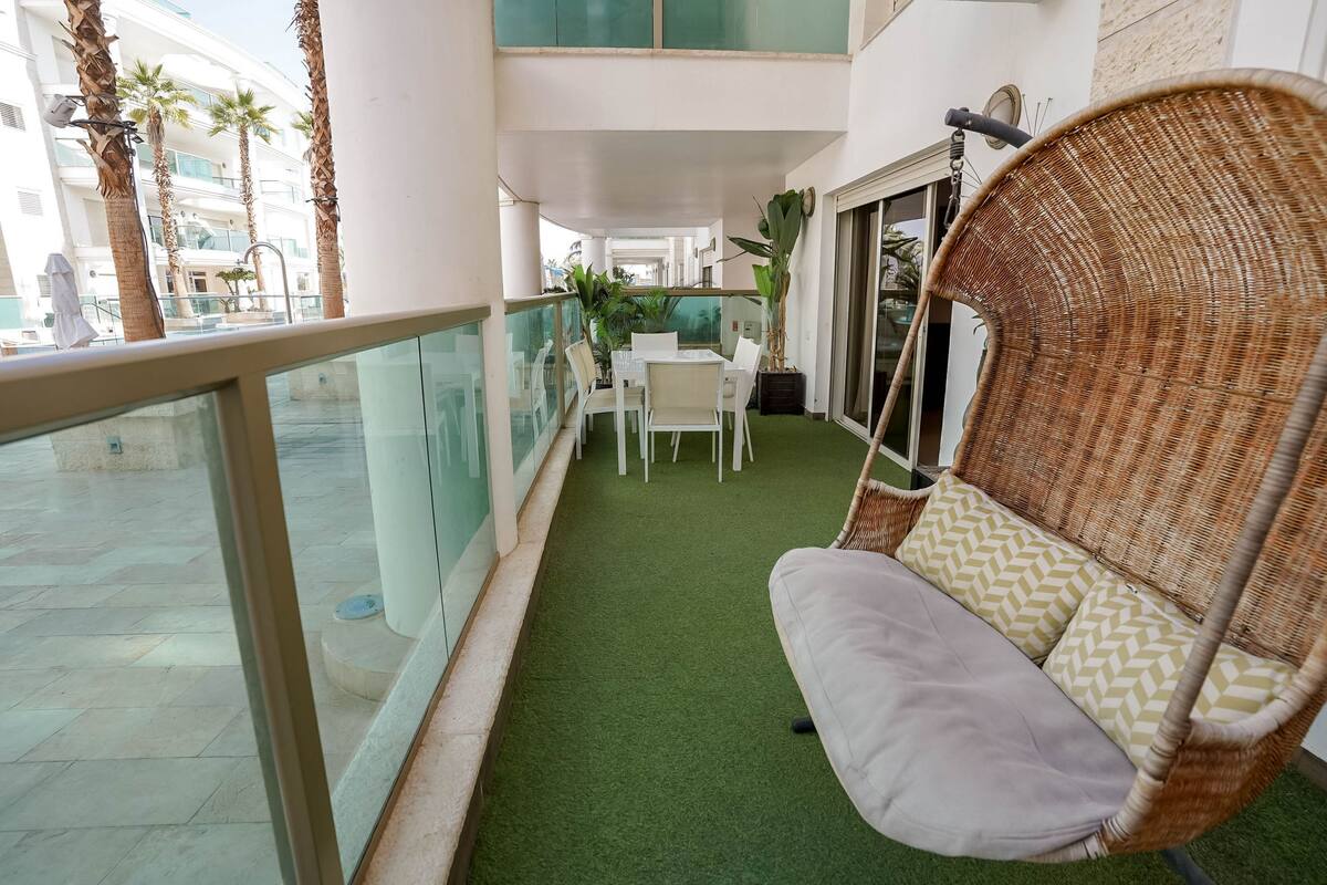 A balcony with a wicker chair, a small table, and chairs, surrounded by potted plants and palm trees.