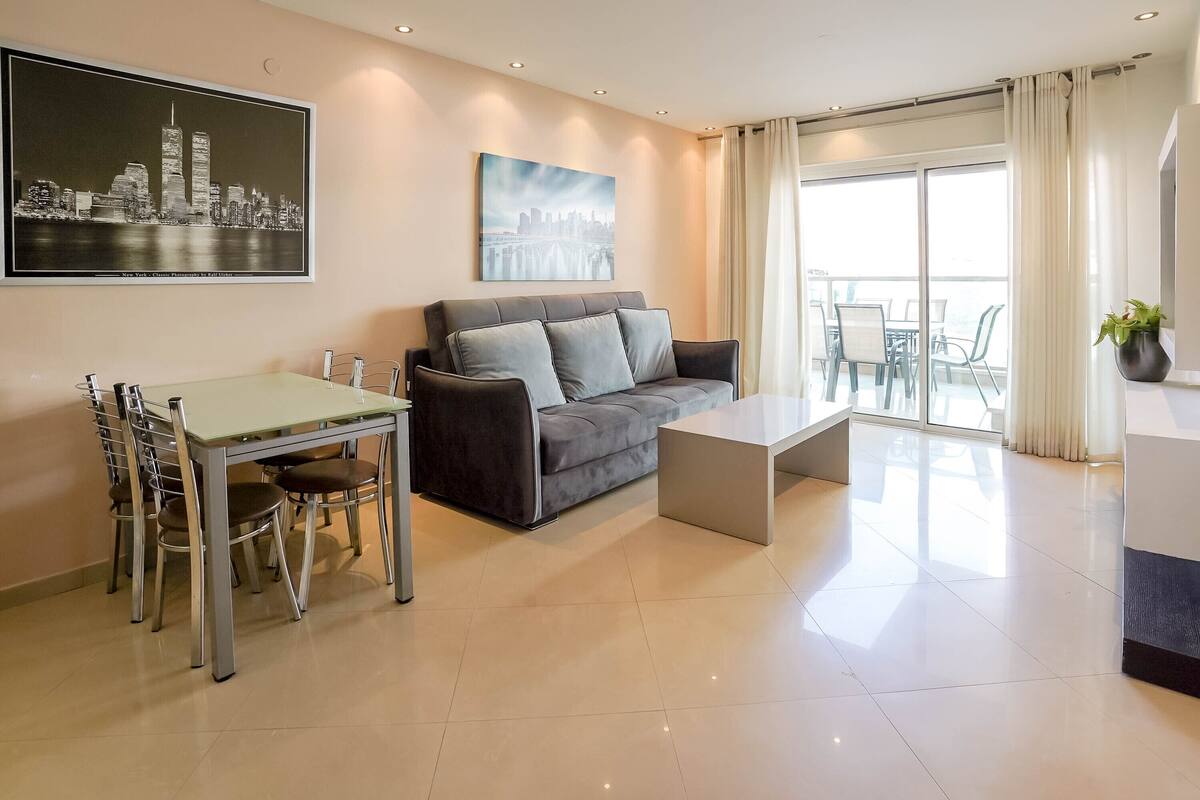 A modern living room with a sofa, a dining table with chairs, a coffee table, and a balcony with a view.