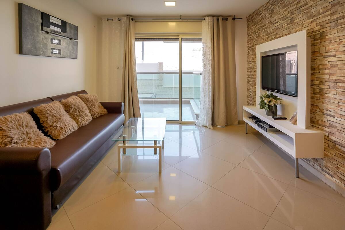 A modern living room with a brown sofa, a glass coffee table, a TV on a white stand, and a stone-patterned wall.