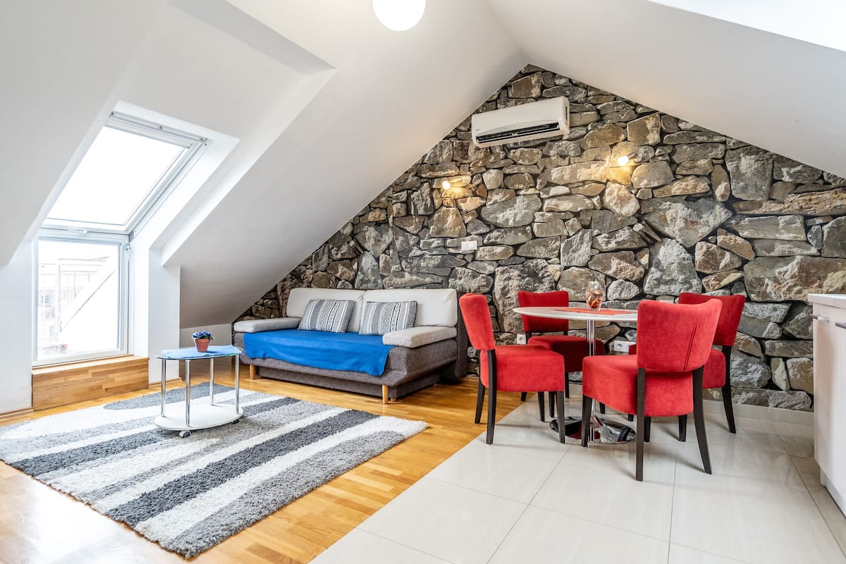 Une pièce de style loft avec un mur en pierre, un coin repas avec des chaises rouges, un canapé agrémenté d’un plaid bleu et une lucarne.