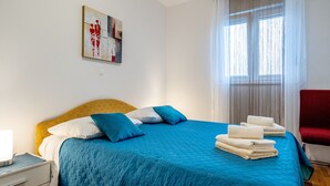 A neatly made bed with blue bedding, a bedside table with a lamp, and a window with sheer curtains. - Apartments Adriatic Split-Stobreč (Split)