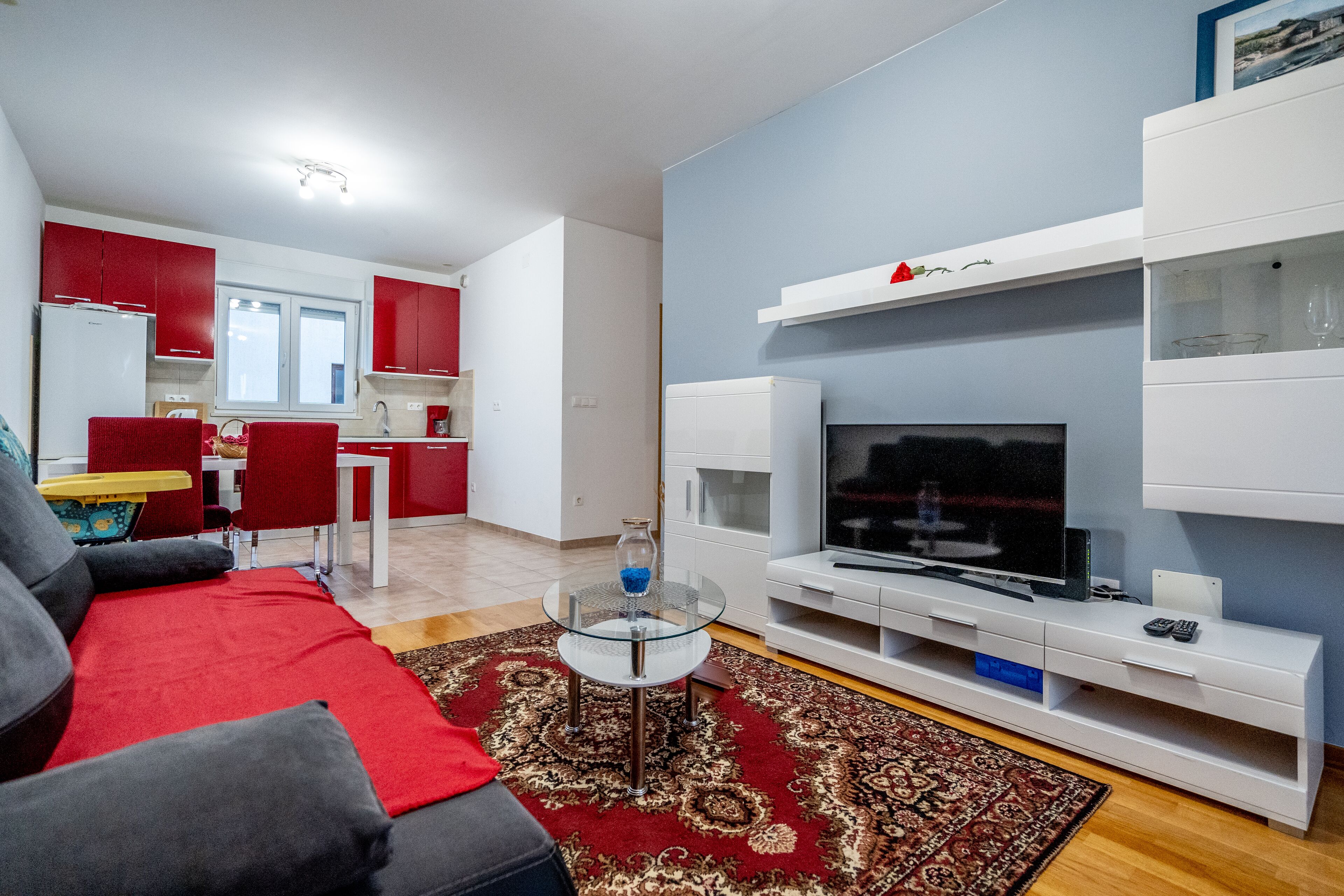 Egy modern nappali, szürke kanapéval, piros párnákkal, üveg kávéasztallal és egy fehér állványon elhelyezett televízióval.