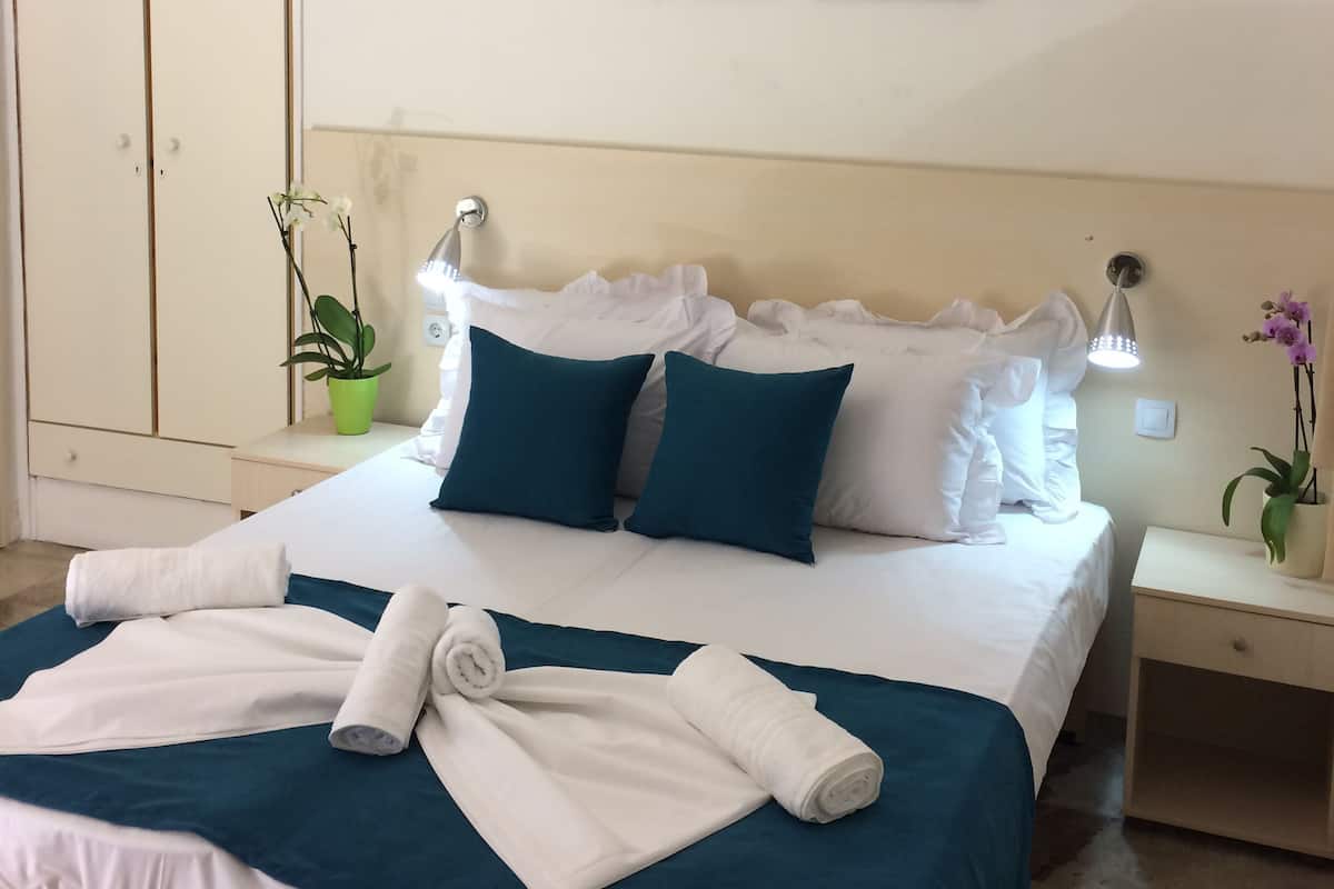 Una cama bien hecha con ropa de cama blanca y verde azulado, dos almohadas verde azulado y un par de toallas blancas enrolladas. Hay dos cuadros de flores en la pared, una lámpara de noche y una pequeña planta en maceta sobre la mesita de noche.
