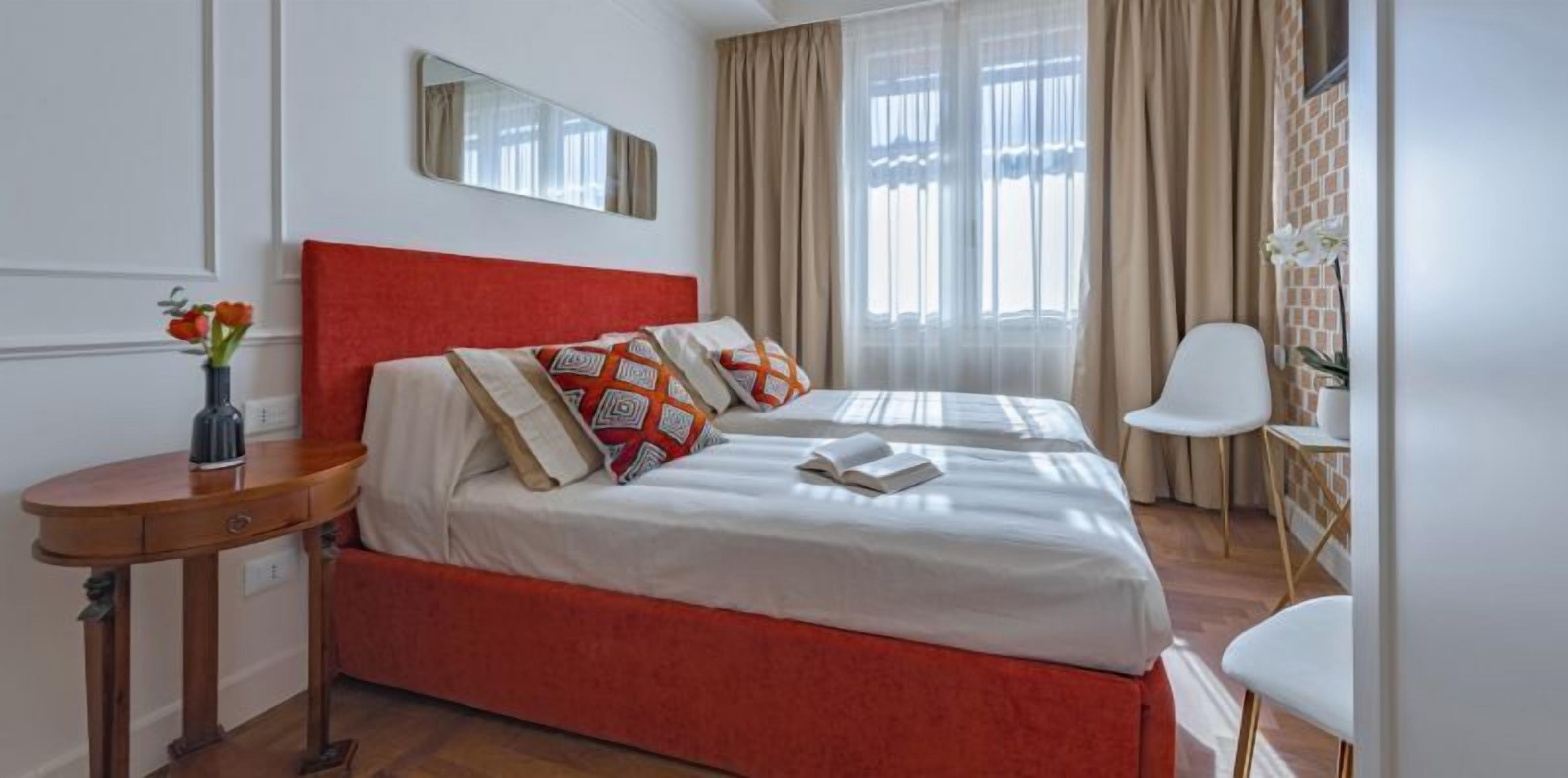 Ein Hotelzimmer mit rotem Kopfteil, einem Bett mit weißer Bettwäsche und gemusterten Kissen, einem Nachttisch mit Lampe und einem Sessel.