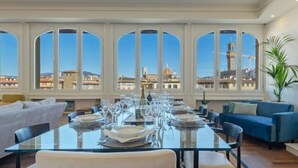 Un área de comedor con una larga mesa dispuesta para una comida, ventanas amplias con vista a la ciudad, un sofá azul y una chimenea.