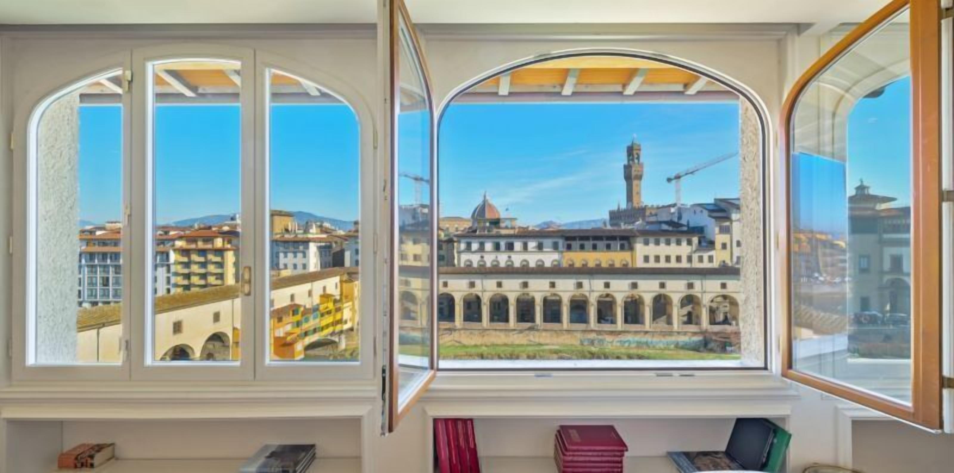 Ein Zimmer mit großen Bogenfenstern und Blick auf eine historische Stadt mit Gebäuden und einem Turm.