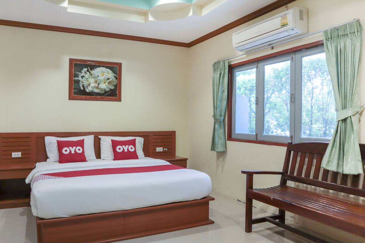 A bedroom with a bed, a bench, a window with curtains, an air conditioning unit, and a framed picture on the wall.