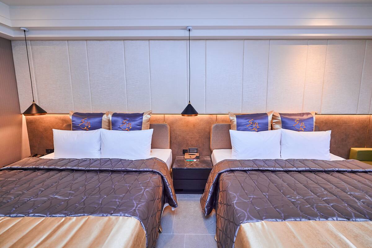 Quarto de hotel com duas camas, uma mesinha de cabeceira e uma luminária na parede.