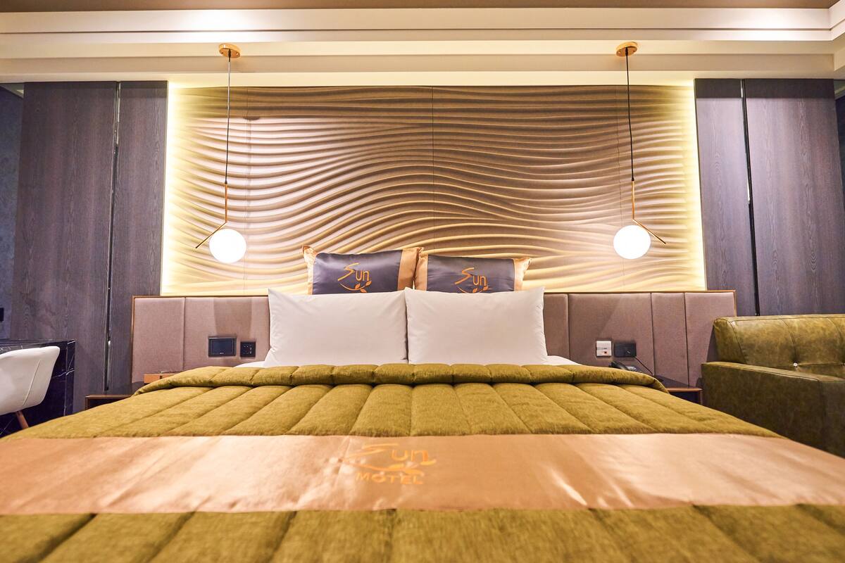 Quarto de hotel moderno com uma cama grande, duas mesas de cabeceira, uma poltrona verde e uma cabeceira com um padrão ondulado único.