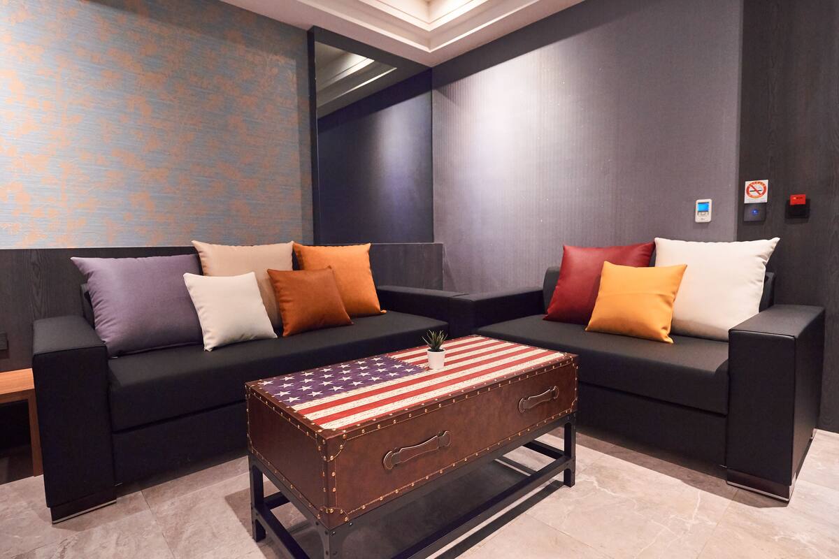 Uma sala de estar moderna com dois sofás pretos, almofadas coloridas e uma mesa de centro em formato de mala antiga com estampa da bandeira americana.