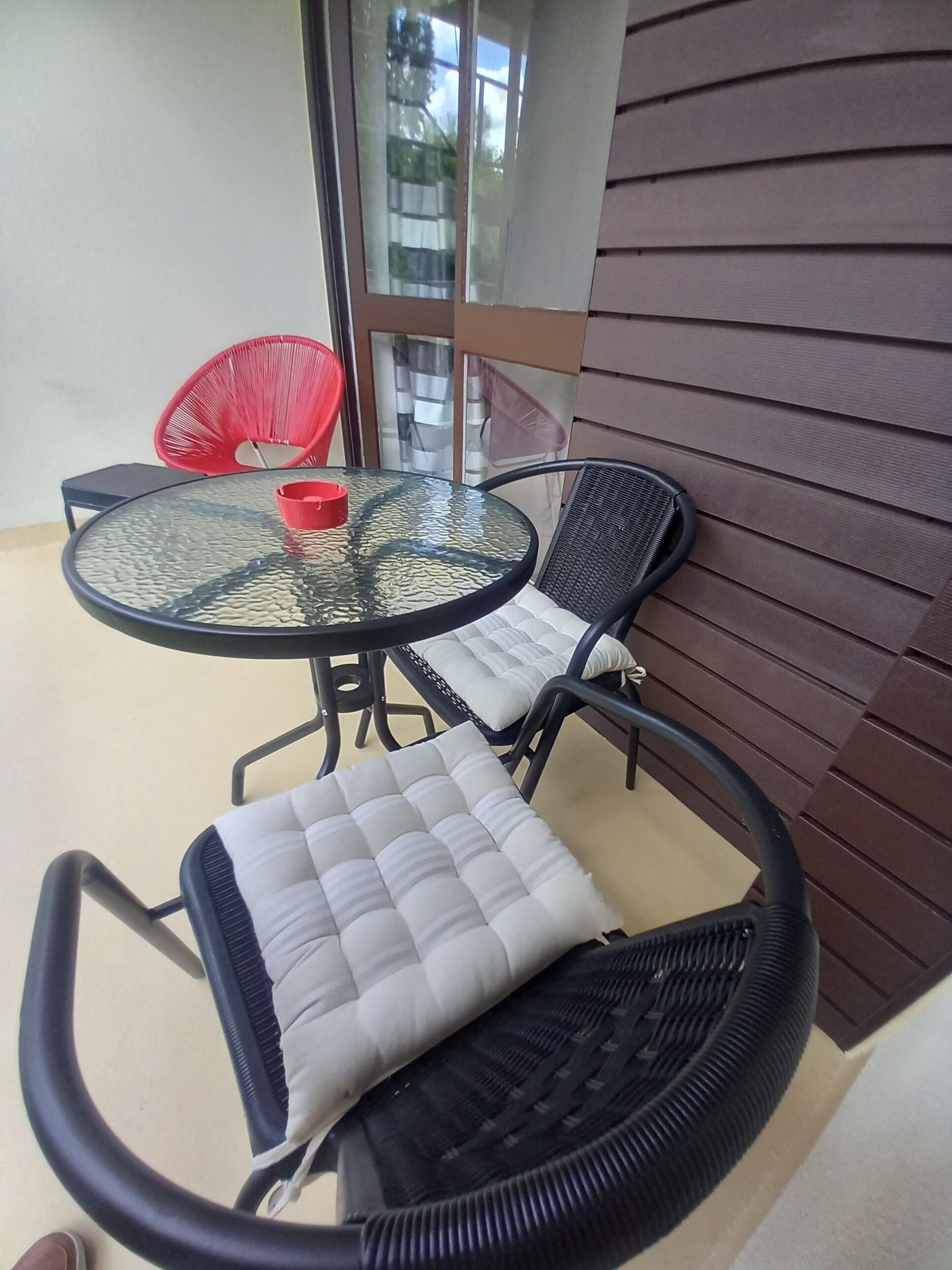 Un pequeño espacio al aire libre con una mesa redonda de vidrio y dos sillas de mimbre negro con cojines blancos.