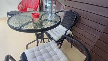 Un pequeño espacio al aire libre con una mesa redonda de vidrio y dos sillas de mimbre negro con cojines blancos.