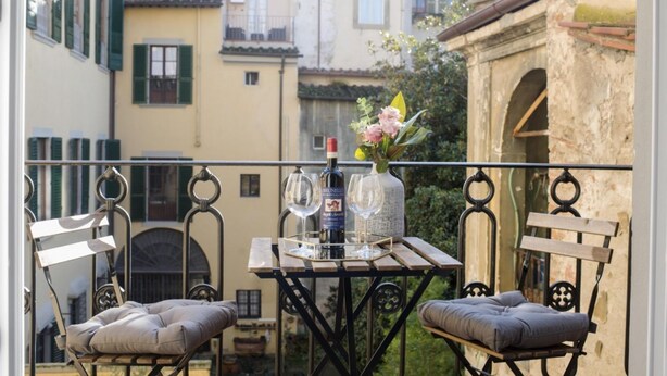 A balcony with two cushioned chairs, a table with a bottle and glasses, and a vase with flowers. - Apartments Florence Oblate Exclusive (Florence)