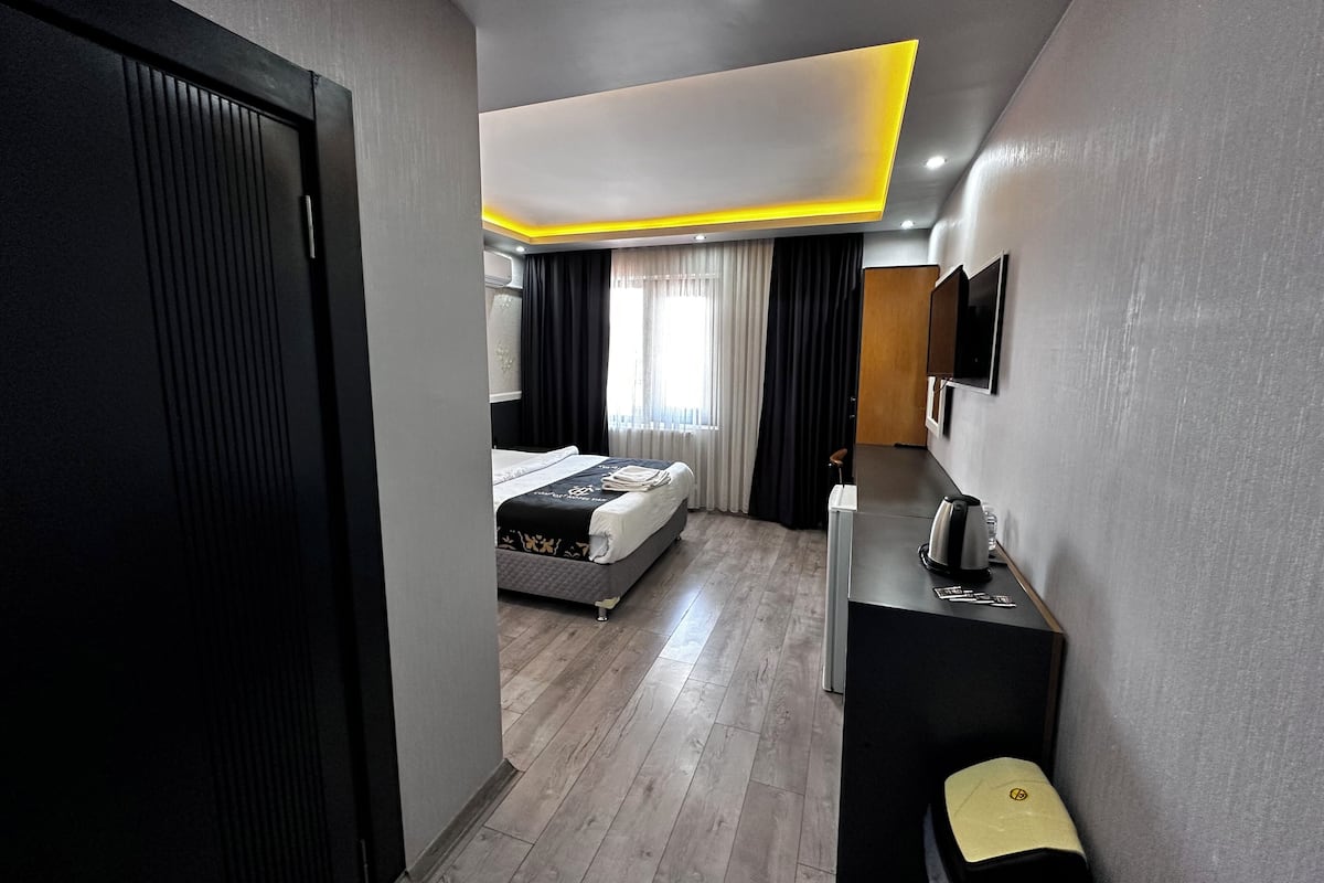 A modern hotel room with a bed, a desk with a kettle, a chair, and a television on the wall.