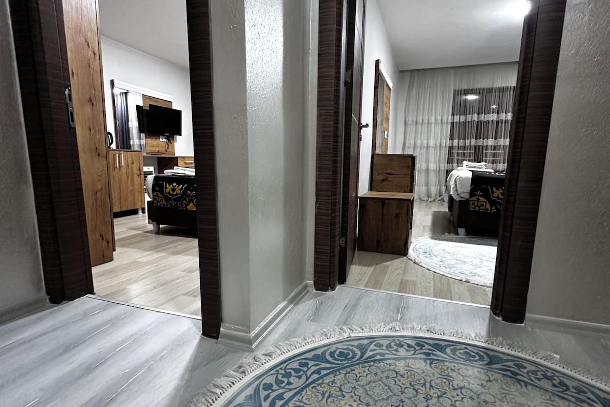 A hallway with a patterned rug, wooden doors, and a view into a bedroom with a bed and curtains.