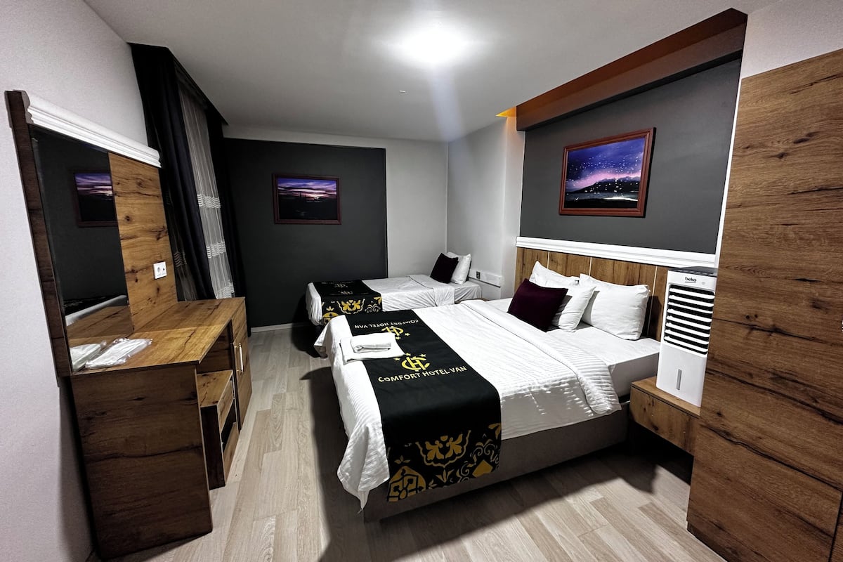 A hotel room with two beds, a desk, and a nightstand. There is a painting on the wall and a Beko air conditioner.