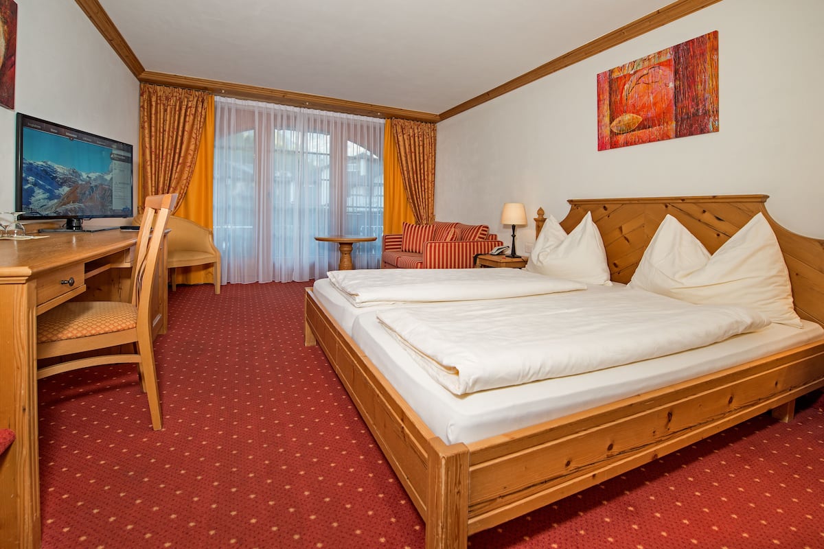A hotel room with a wooden bed, a desk with a computer, a chair, a small table, and a window with curtains.