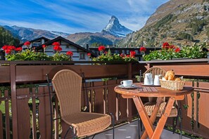 A balcony with wicker furniture, a table with breakfast items, and a view of a mountain with red flowers in the foreground. - Hotel Dufour Chalet (Zermatt)