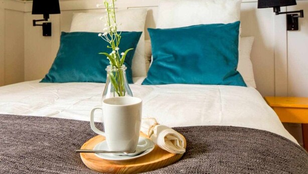 A neatly made bed with white and teal pillows, a glass vase with flowers, a white mug, and a folded napkin on a wooden coaster.