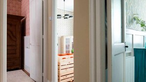 A hallway with a wooden door, a white door, and a view into a room with a wooden dresser and a hanging light fixture.
