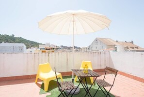 Een dakterras met een tafel en stoelen, een gele plastic stoel, een houten tafel en een beige parasol.