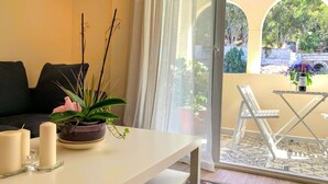 Una sala de estar con un sofá, una mesa con velas y una planta, un balcón con mesa y sillas, y vistas a zonas verdes y un puente.