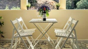 Un pequeño espacio al aire libre con dos sillas blancas y una mesa, una botella de vino y un jarrón con flores púrpuras.