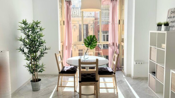 Een eetgedeelte met een ronde tafel, stoelen, een potplant en uitzicht op gebouwen door een raam met roze gordijnen.