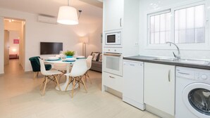 Una cocina moderna con armarios blancos, horno empotrado, microondas y lavavajillas. Cuenta con una zona de comedor con mesa y sillas, y un televisor montado en la pared.