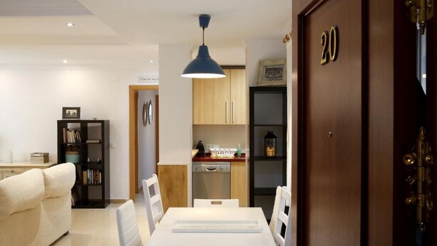 Un soggiorno moderno con un divano bianco, un tavolo da pranzo con sedie, un angolo cottura con mobili in legno e una lampada a sospensione blu.