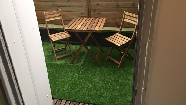Un petit espace extérieur aménagé avec une table en bois et deux chaises, entouré d’une clôture en bois et doté d’un sol recouvert d’un gazon artificiel vert.