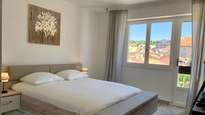 A bedroom with a large bed, a nightstand, a wall-mounted lamp, and a window with a view of buildings. - 4-person apartment facing the sea 50m from the beach and town center (Sainte-Maxime)