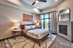 A bedroom with a bed, two bedside tables, a fireplace, a large window with curtains, and a ceiling fan.
