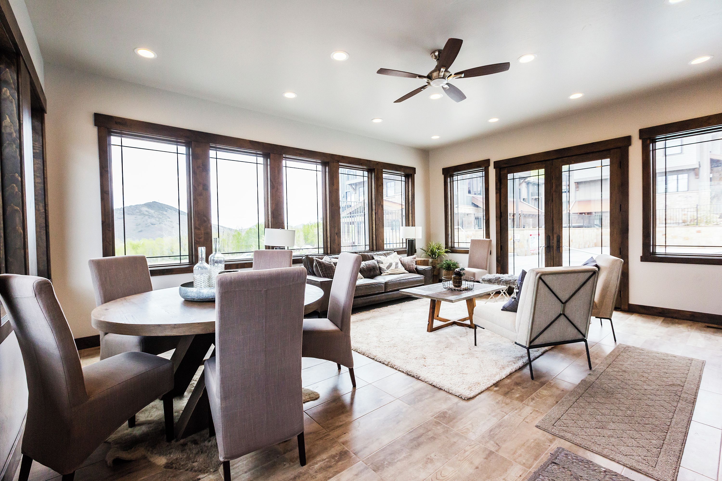 A spacious living room with a round dining table, grey upholstered chairs, a sofa, and a coffee table. There are large windows providing natural light and views of the outside.