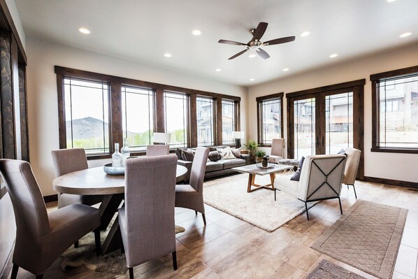 A spacious living room with a round dining table, grey upholstered chairs, a sofa, and a coffee table. There are large windows providing natural light and views of the outside.