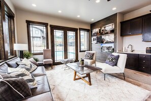 Una sala de estar moderna con sofá, mesa de centro y sillón, una zona de cocina con armarios oscuros y ventanas grandes que ofrecen vistas al exterior.