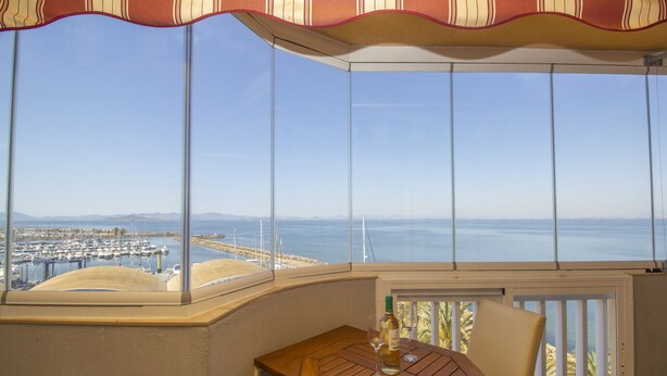 Un balcón en esquina con una mesa y una silla de madera, con vista a un puerto deportivo y al mar.