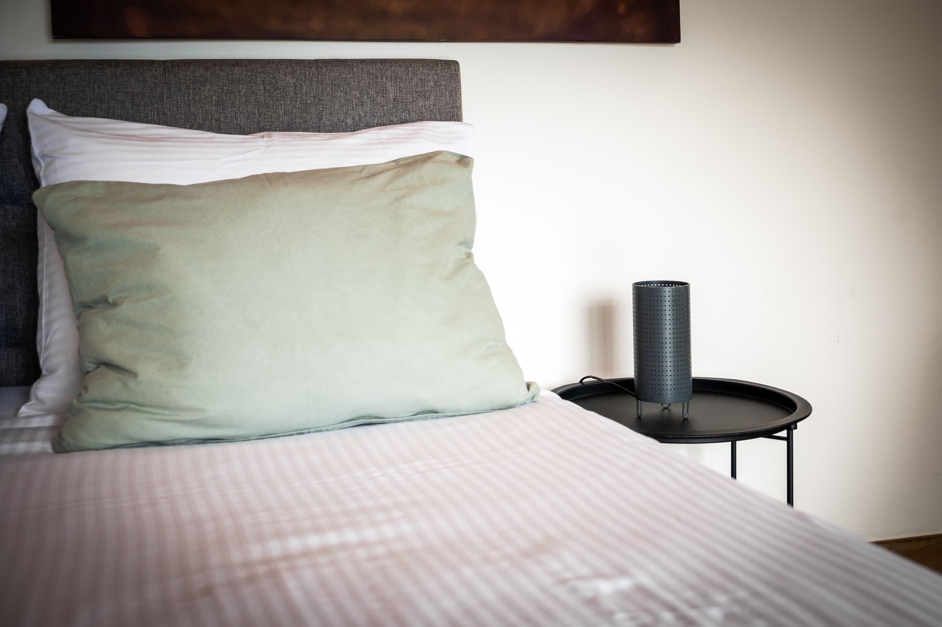 A neatly made bed with a striped bedspread and two pillows, a small black side table with a cylindrical object on it, and a plain wall with a dark artwork above the bed.