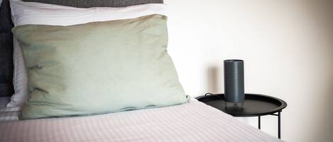 A neatly made bed with a striped bedspread and two pillows, a small black side table with a cylindrical object on it, and a plain wall with a dark artwork above the bed.