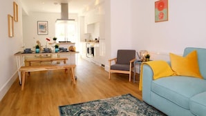 A modern living room with a blue sofa, wooden dining table, and a kitchen in the background. - NEW Poole House 5 mins to Sandbanks +Netflix (Poole)