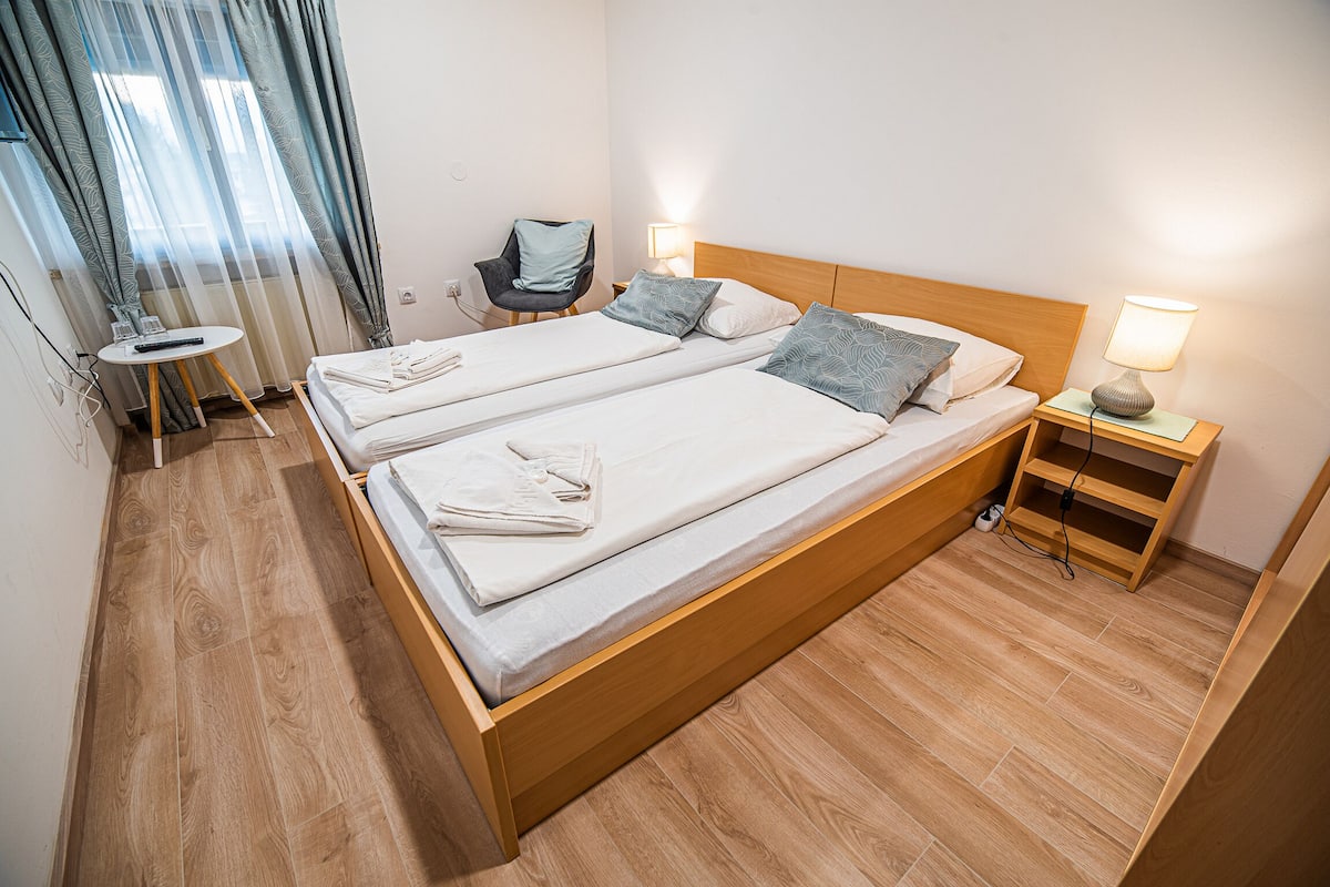Cama doble con dos almohadas, mesita de noche con lámpara, una silla y una pequeña mesa con control remoto.