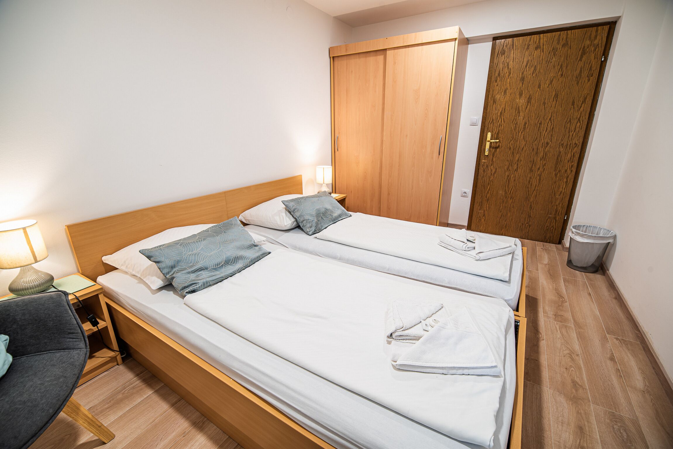 Habitación de hotel con dos camas individuales, un armario de madera, un escritorio con lámpara y una silla.