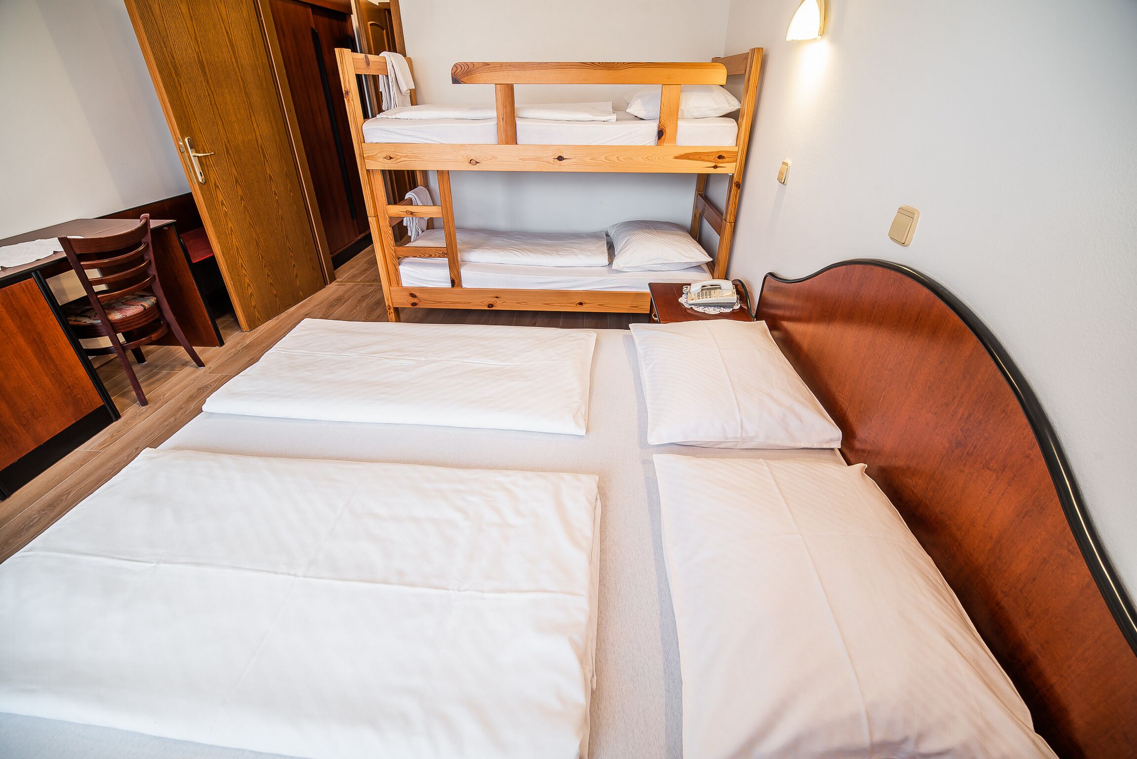 Una cama litera de dos plazas, un escritorio de madera con una silla y una mesita de noche con una lámpara.
